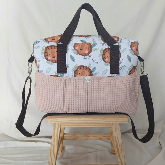 Vista frontal de la bolsa de maternidad, de tejido estampado de leonas en fondo blanco, con bolsillos de tela de waffle rosada en el tercio inferior. Asas de cinta plana negra de 3 cm de ancho y cinta idéntica para colgar al cuello en la parte superior.