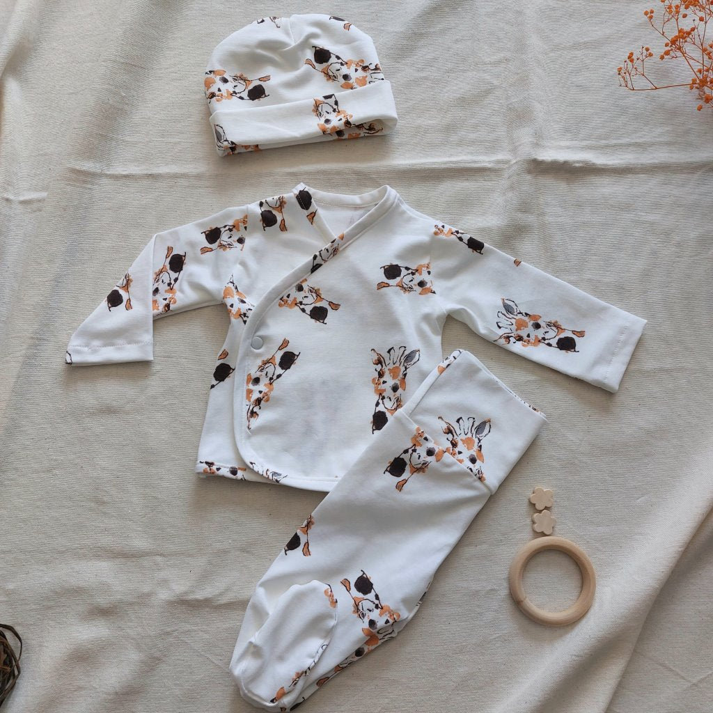 Conjunto de recién nacido, compuesto por una camisa con cierre en el lateral de la barriga, camisa, polainas y gorrito con estampado de pequeñas cabezas de jirafa de topos chocolate y marrón caramelo sobre fondo blanco.