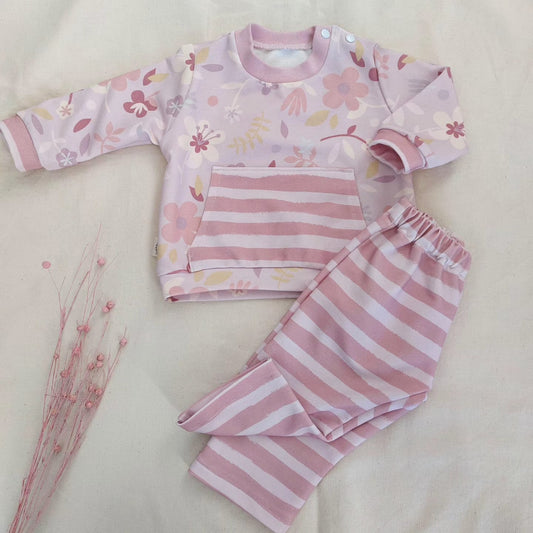Vista frontal de conjunto de bebé que incluye una sudadera con estampado de flores sobre fondo rosado, con un práctico bolsillo a dos manos. Los pantalones se ven doblados por la mitad y muestran rayas de colores rosa y blanco.