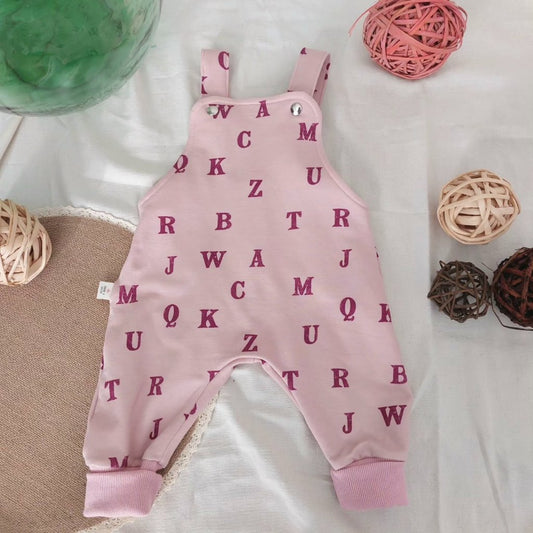 Peto para bebé, visto de frente con estampado de letras lilas en fondo rosa con puños de tobillo también rosa