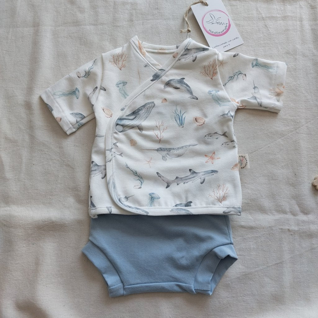 Conjunto de recién nacido, compuesto por camisa de manga corta con cierre en el lateral de la barriga, estampado de fauna marina en fondo blanco. Completado con pantaloncitos cortos de color celeste.