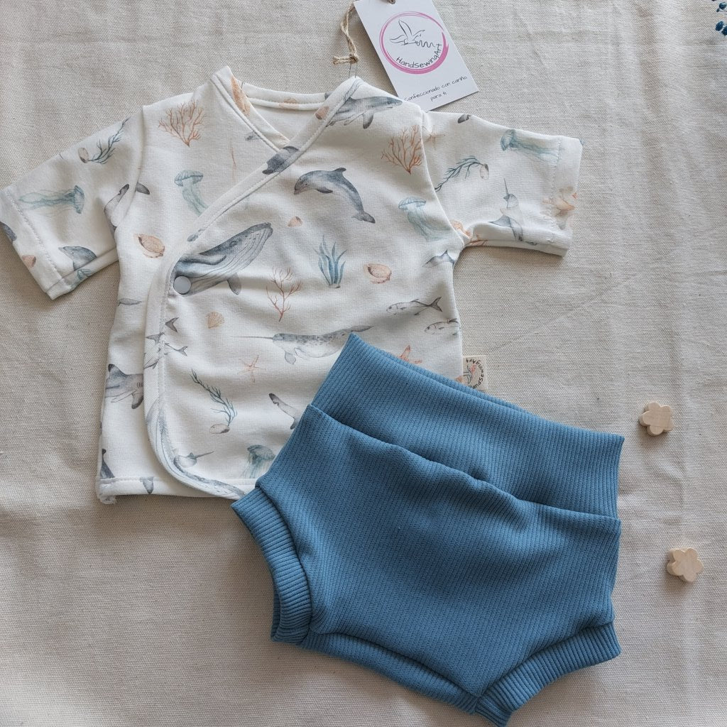 Conjunto de recién nacido, compuesto por camisa de manga corta con cierre en el lateral de la barriga, estampado de fauna marina en fondo blanco. Completado con pantaloncitos cortos de color azul colocados en diagonal.