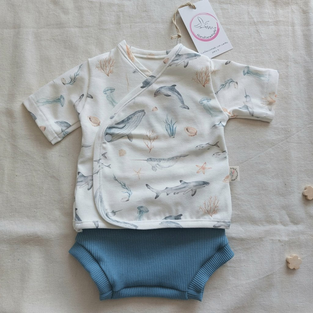 Conjunto de recién nacido, compuesto por camisa de manga corta con cierre en el lateral de la barriga, estampado de fauna marina en fondo blanco. Completado con pantaloncitos cortos de color azul.