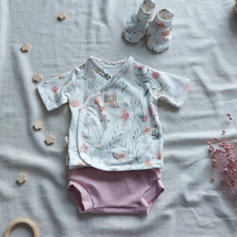 Conjunto de recién nacido, compuesto por camisa de manga corta con cierre en el lateral de la barriga, estampado de mariposas con flores. Completado con pantaloncitos cortos de color rosa en la parte inferior y de patucos estampados de mariposas en la parte superior derecha.