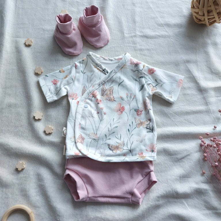 Conjunto de recién nacido, compuesto por camisa de manga corta con cierre en el lateral de la barriga, estampado de mariposas con flores. Completado con pantaloncitos cortos de color rosa en la parte inferior y de patucos de color rosa en la parte superior izquierda.