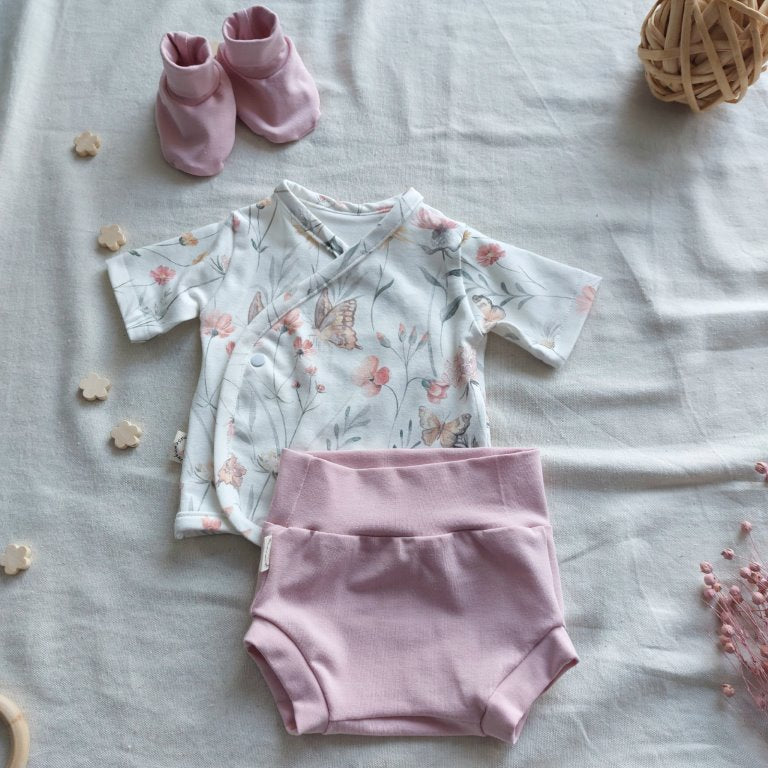 Conjunto de recién nacido, compuesto por camisa de manga corta con cierre en el lateral de la barriga, estampado de mariposas con flores. Completado con pantaloncitos cortos de color rosa en la parte inferior superpuestos en la camisa y de patucos de color rosa en la parte superior izquierda.