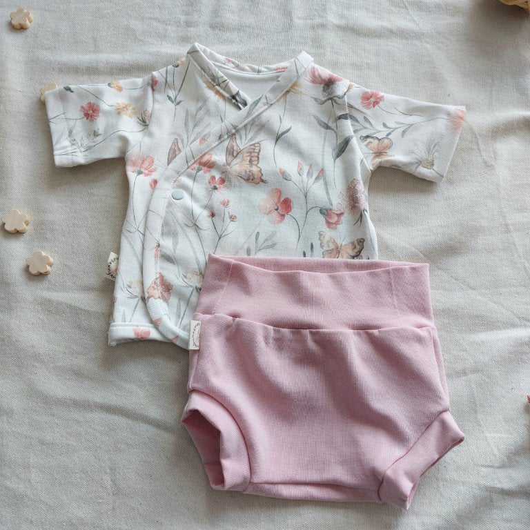 Conjunto de recién nacido, compuesto por camisa de manga corta con cierre en el lateral de la barriga, estampado de mariposas con flores. Completado con pantaloncitos cortos rosas sobrepuestos sobre la parte inferior izquierda de la camisa.