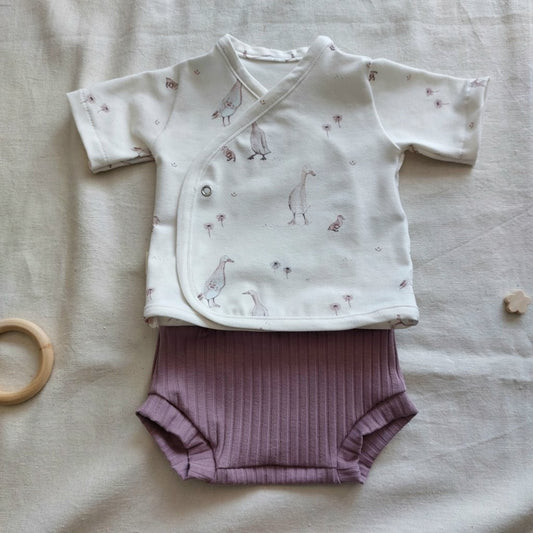 Conjunto de recién nacido, compuesto por camisa de manga corta con cierre en el lateral de la barriga, con estampado de ocas sobre fondo blanco. Completado con pantaloncitos cortos de color lila en la parte inferior.