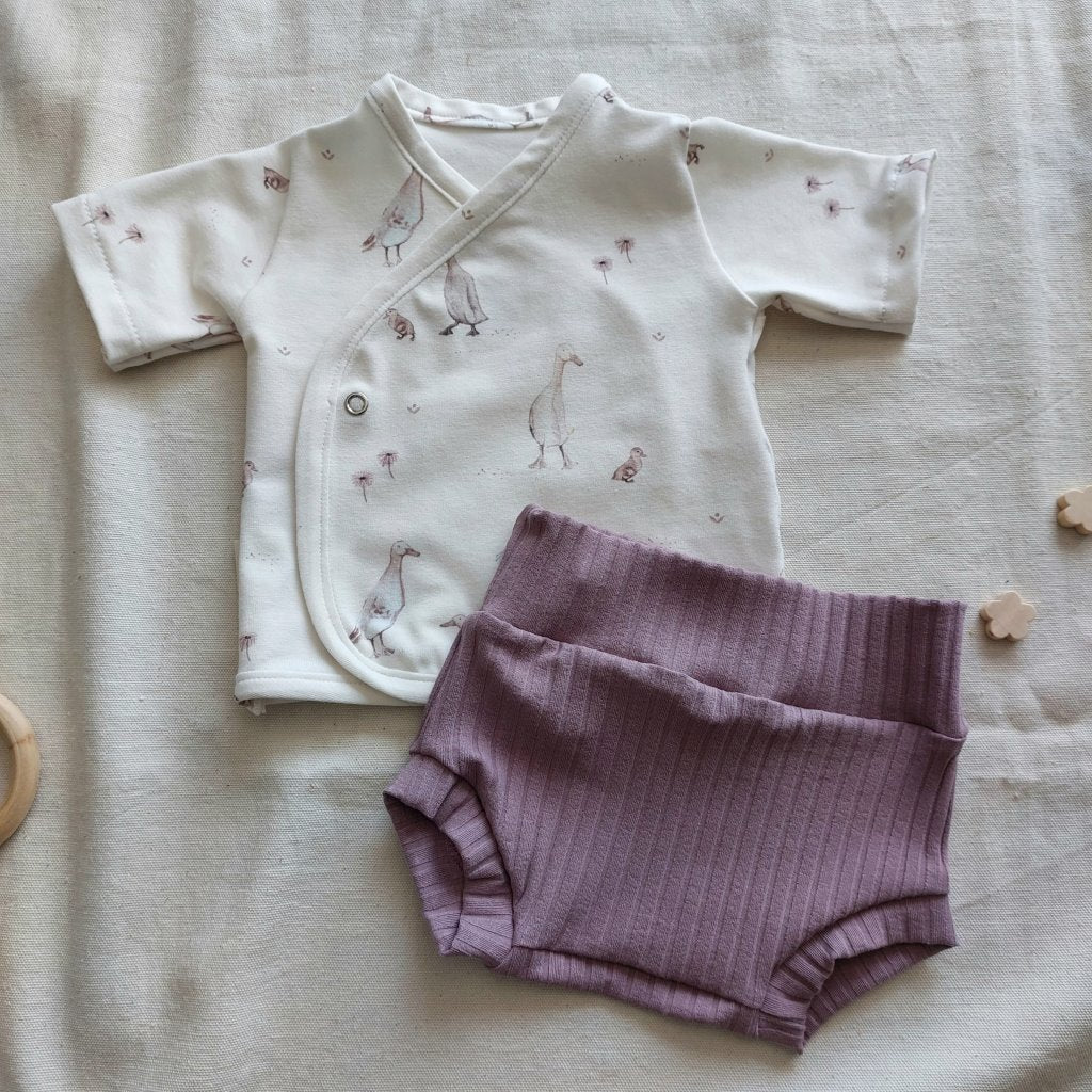 Conjunto de recién nacido, compuesto por camisa de manga corta con cierre en el lateral de la barriga, con estampado de ocas sobre fondo blanco. Completado con pantaloncitos cortos de color lila colocados en diagonal.